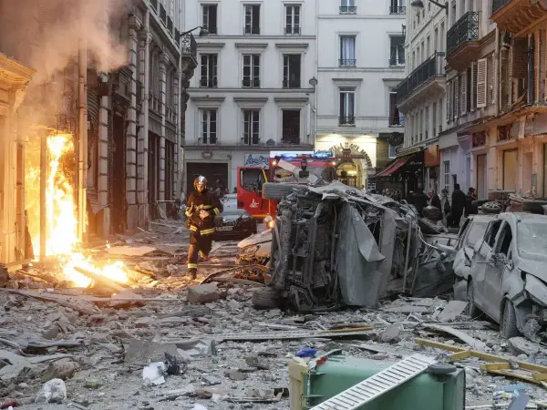 Un pompier avance dans les décombres d'une explosion gaz Paris. Des voitures détruites et des flammes ravagent la rue.